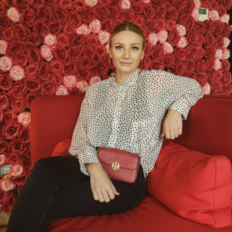 Junge Frau auf einem roten Sofa mit schwarzer Hose und weisser Bluse mit Rosenbild im Hintergrund