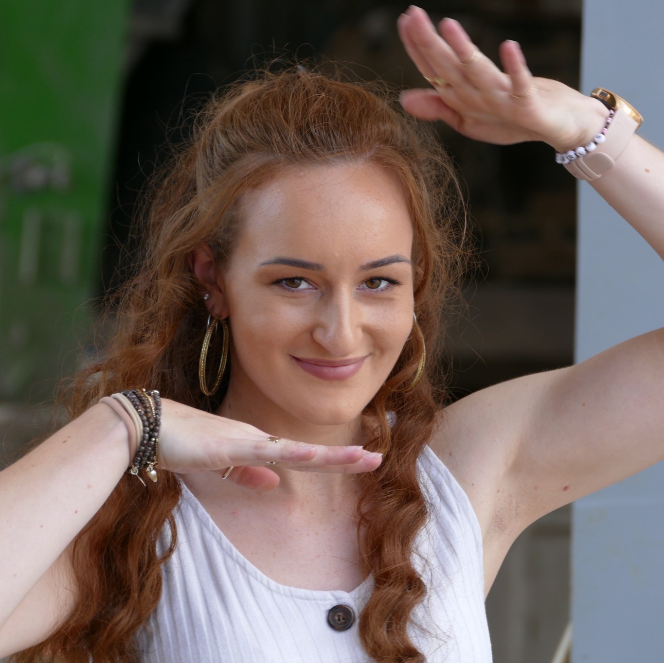 Frau mit rötlichen langen gewellten Haaren steht draussen und trägt ein weissen Top. Sie hat die Arme zu einem Foto geformt und lächelt